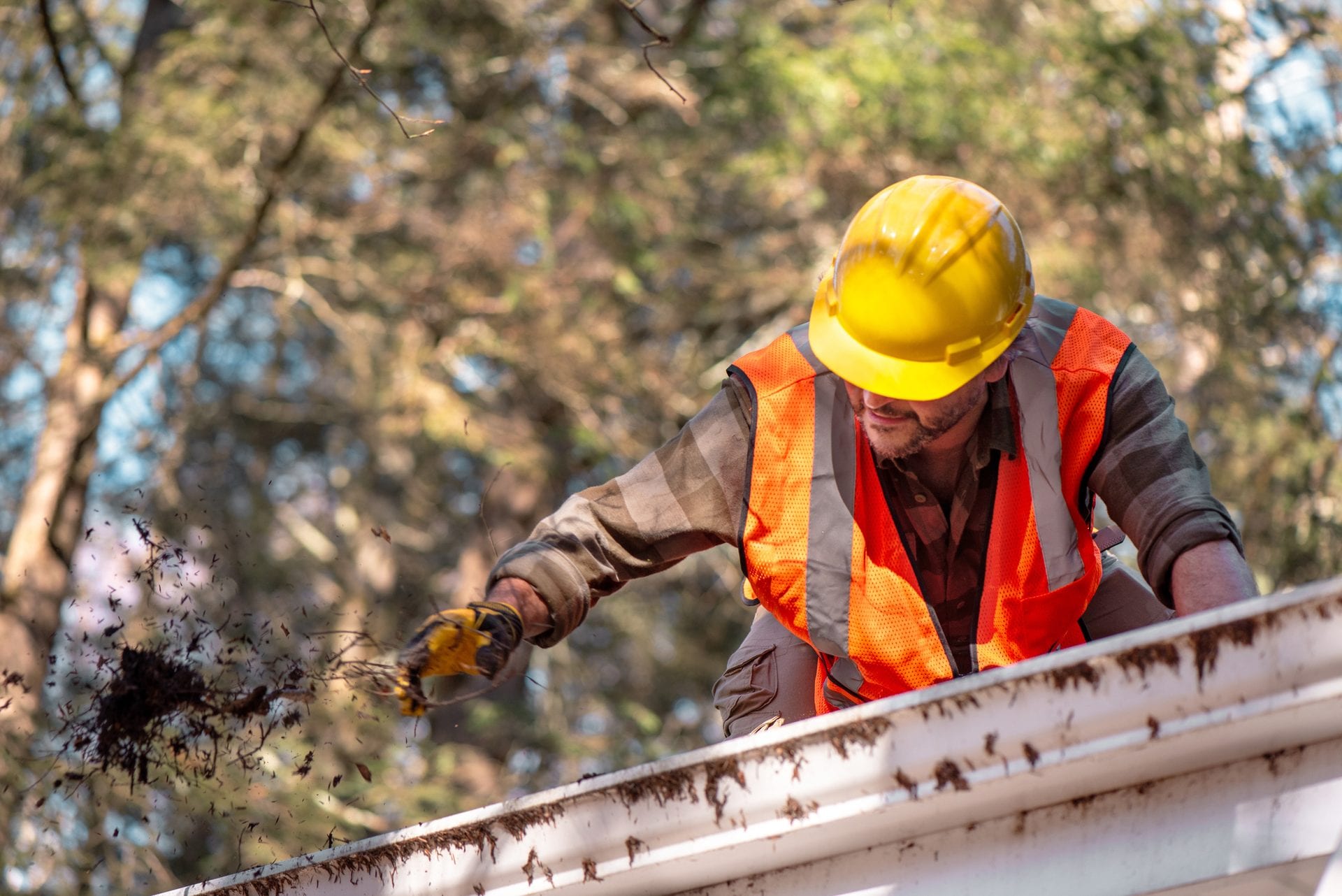 A worker in a hard hat and high visibility vest scooping leaf debris out of a roof gutter